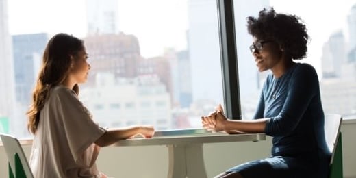 two women chatting at the table next to a window.
